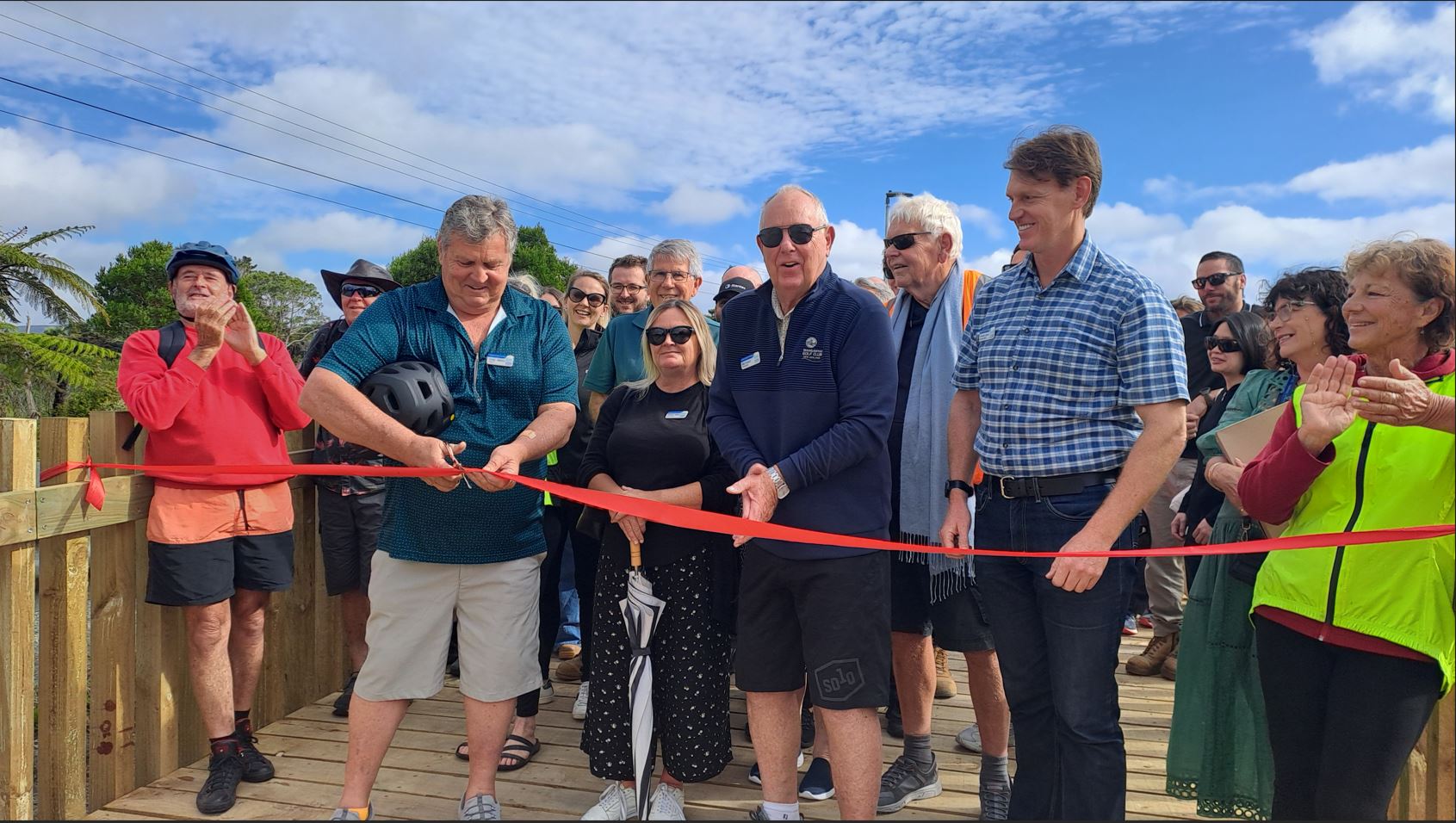 A group of people stand in front of a red ribbon tied across a wooden boardwalk. A man with a bike helmet under his arm and a pair of scissors poised to cut the red ribbon. 